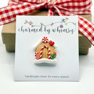 May include: A gingerbread house pin on a white card with the text "charmed by whimsy" and "handmade cheer for every season." The pin is decorated with candy canes, a red heart, and green and red candies. The card is placed in front of a brown gift box tied with a red and white checkered ribbon.