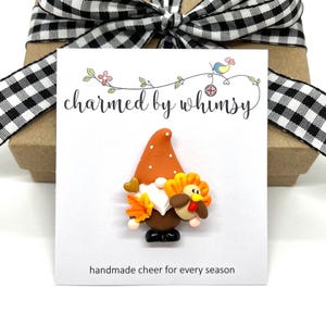 May include: A small, handmade gnome figurine with a brown hat and white beard, holding a small turkey figurine. The gnome is wearing black shoes and has a brown body. The figurine is on a white card with the text "charmed by whimsy" and "handmade cheer for every season".