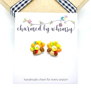 May include: A pair of turkey-shaped stud earrings on a white display card. The earrings feature a tan body, a yellow beak, a red wattle, and a multi-colored fan-shaped tail in yellow, orange, and green. The card displays the text charmed by whimsy.