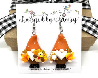 May include: A pair of whimsical gnome earrings. Each earring features a gnome with an orange hat with white dots, a white beard, and a brown body. The gnomes hold a turkey and fall leaves. The card reads "charmed by whimsy".