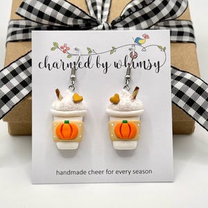 May include: A pair of dangle earrings shaped like coffee cups with white whipped cream, a small brown heart, and a tan sleeve featuring an orange pumpkin graphic. The earrings are displayed on a white card with the text charmed by whimsy.