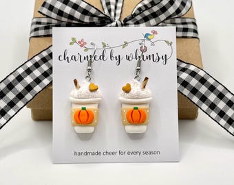 May include: A pair of dangle earrings shaped like coffee cups with white whipped cream, a small brown heart, and a tan sleeve featuring an orange pumpkin graphic. The earrings are displayed on a white card with the text charmed by whimsy.