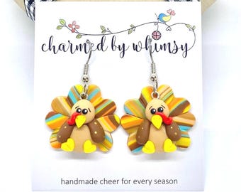 May include: A pair of turkey-shaped earrings with colorful striped tail feathers, brown bodies, and yellow feet. The earrings are on a white card with the text "charmed by whimsy" and "handmade cheer for every season".
