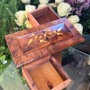 Peut inclure: Boîte à bijoux en bois de forme rectangulaire avec couvercle. Le bois est brun foncé avec une incrustation décorative en bois clair, motif floral. La boîte est ouverte, révélant l'intérieur.