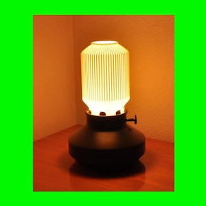 May include: A black table lamp with a cylindrical, ribbed, off-white shade. The lamp has a rounded base and a small, black knob. The lamp is lit, emitting a warm, yellow glow. The lamp is on a wooden surface.