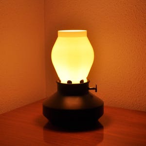 May include: A vintage-style table lamp with a black base and a white, rounded glass shade. The lamp is illuminated, emitting a warm, yellow glow. The lamp is on a wooden surface.