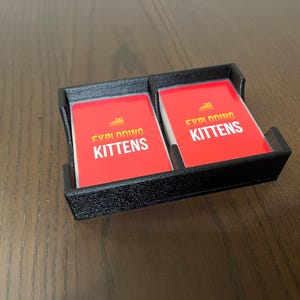 May include: A black plastic card holder with two red cards that say "Exploding Kittens" in yellow letters.