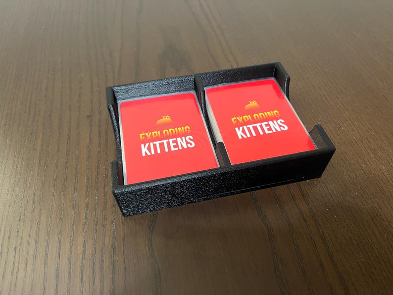 May include: A black plastic card holder with two red cards that say "Exploding Kittens" in yellow letters.