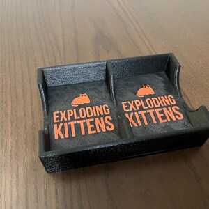 May include: A black plastic card holder with two compartments. Each compartment has a black and white card with the text "Exploding Kittens" and a cartoon orange cat.