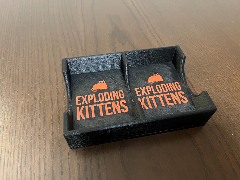 May include: A black plastic card holder with two compartments. Each compartment has a black and white card with the text "Exploding Kittens" and a cartoon orange cat.