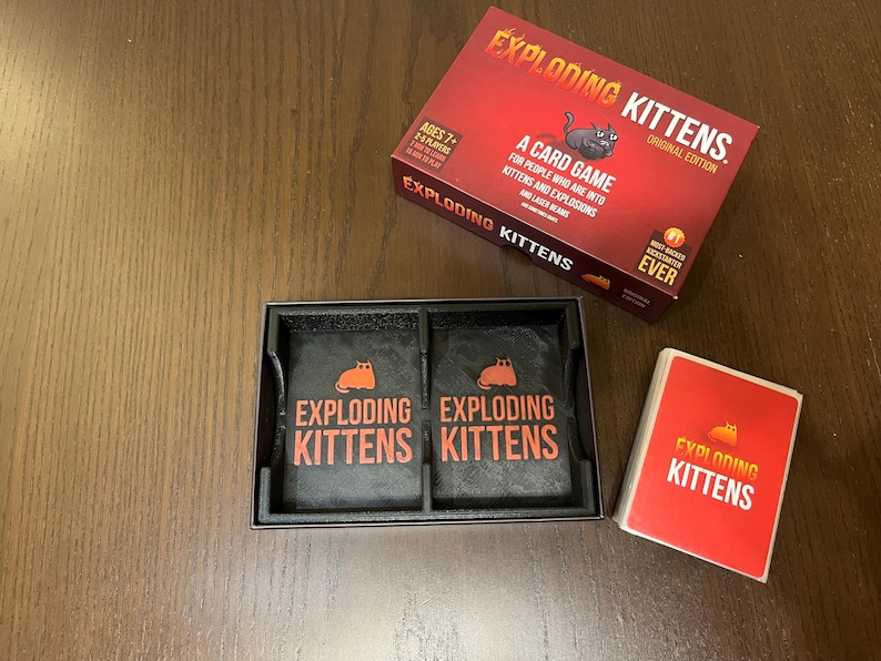 May include: A red box with the words "Exploding Kittens" on it. The box is open and shows two black card holders with the words "Exploding Kittens" on them. There is also a red card with the words "Exploding Kittens" on it.