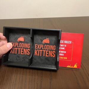 May include: A black card game holder with two compartments, each labeled "Exploding Kittens". The holder is made of plastic and has a textured surface. A red card with the text "These rules!" is visible in the background.