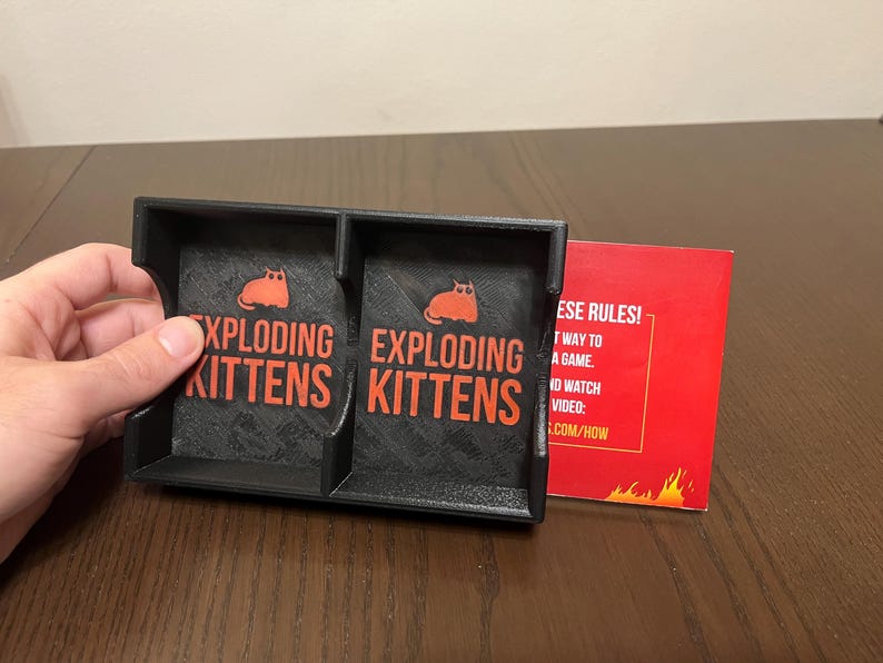 May include: A black card game holder with two compartments, each labeled "Exploding Kittens". The holder is made of plastic and has a textured surface. A red card with the text "These rules!" is visible in the background.