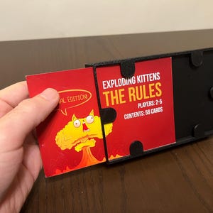 May include: A black card game box with a red card that says "Exploding Kittens The Rules" and "Players: 2-5" and "Contents: 56 Cards". The card is partially pulled out of the box. The box has a black lid with a red card that says "Exploding Kittens" and a cartoon cat with a bomb.