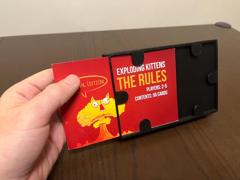 May include: A black card game box with a red card that says "Exploding Kittens The Rules" and "Players: 2-5" and "Contents: 56 Cards". The card is partially pulled out of the box. The box has a black lid with a red card that says "Exploding Kittens" and a cartoon cat with a bomb.