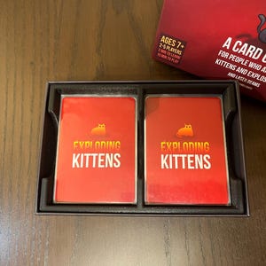 May include: Two red Exploding Kittens playing cards in a black box. The cards have a yellow cat with a bomb on its back and the text "Exploding Kittens".
