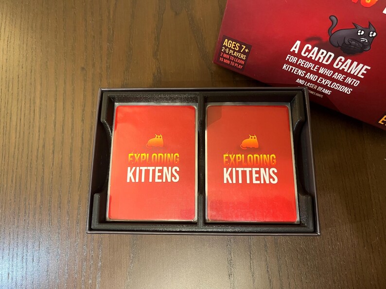 May include: Two red Exploding Kittens playing cards in a black box. The cards have a yellow cat with a bomb on its back and the text "Exploding Kittens".