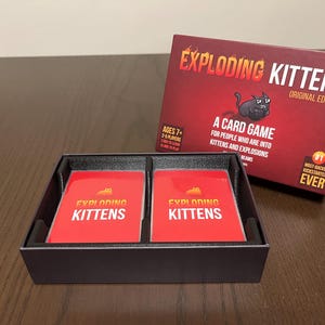 May include: A black box with two red cards inside. The cards have the words "Exploding Kittens" printed on them. The box is labeled "Exploding Kittens Original Edition".