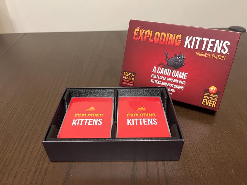 May include: A black box with two red cards inside. The cards have the words "Exploding Kittens" printed on them. The box is labeled "Exploding Kittens Original Edition".