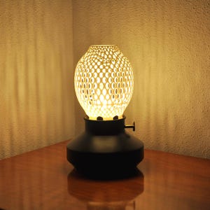 Könnte beinhalten: Eine dekorative Tischlampe mit schwarzem Sockel und weißem, gemustertem Schirm. Der Schirm hat ein einzigartiges, offenes Design, das Licht durchscheinen lässt. Die Lampe steht auf einer Holzoberfläche, der Hintergrund ist eine strukturierte Wand.