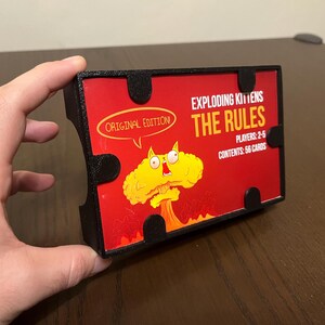 May include: A black card game box with a red and yellow illustration of a cat with a bomb for a head. The text on the box reads "Exploding Kittens The Rules", "Original Edition", "Players: 2-5", and "Contents: 56 Cards".