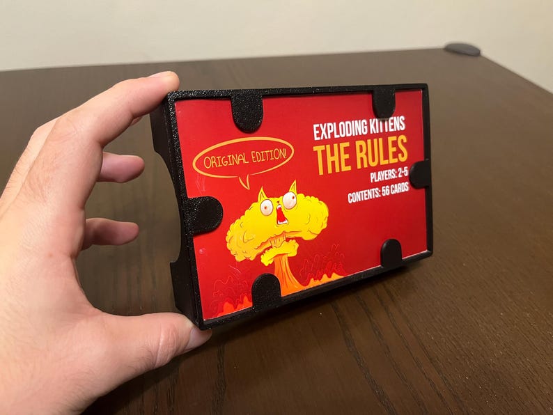 May include: A black card game box with a red and yellow illustration of a cat with a bomb for a head. The text on the box reads "Exploding Kittens The Rules", "Original Edition", "Players: 2-5", and "Contents: 56 Cards".