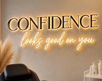 May include: A wall-mounted sign featuring the word CONFIDENCE in large, dark, backlit capital letters, positioned above the phrase looks good on you written in a glowing, cursive neon-style script with a warm, soft yellow illumination.