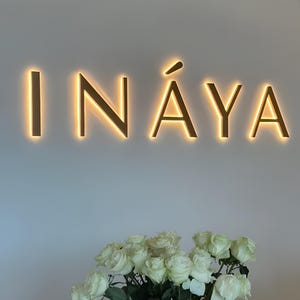以下が含まれることがあります： 白い壁に、金色の照明付き文字で「INAYA」と書かれています。その下に、白いバラのブーケが白い表面に置かれています。全体的な雰囲気はクリーンでモダンで、ビジネスや専門的な環境を示唆しています。
