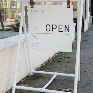 Pu&ograve; includere: Un cartello a cavalletto in metallo bianco con un cartello rettangolare bianco che recita "fig. a" e "OPEN" con una freccia che punta a sinistra. Il cartello &egrave; tenuto da cinghie di cuoio marrone. L'handle di Instagram e il sito web del negozio sono elencati in basso.