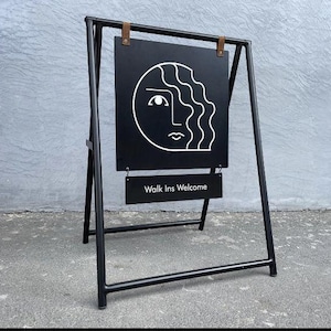 Pu&ograve; includere: Cartello a cavalletto in metallo nero con un design minimalista di un volto bianco. Il cartello mostra il testo "Walk Ins Welcome" in bianco su un cartello nero pi&ugrave; piccolo. Il cartello &egrave; su una superficie di cemento grigio.