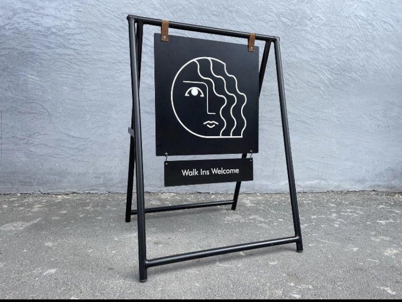 Pu&ograve; includere: Cartello a cavalletto in metallo nero con un design minimalista di un volto bianco. Il cartello mostra il testo "Walk Ins Welcome" in bianco su un cartello nero pi&ugrave; piccolo. Il cartello &egrave; su una superficie di cemento grigio.