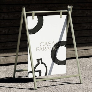 Pu&ograve; includere: Un cavalletto da esterno verde chiaro con un poster bianco. Il poster mostra il testo "CASA PARAMO" e disegni astratti neri. Il cavalletto &egrave; su una superficie grigia, con uno sfondo in legno scuro.