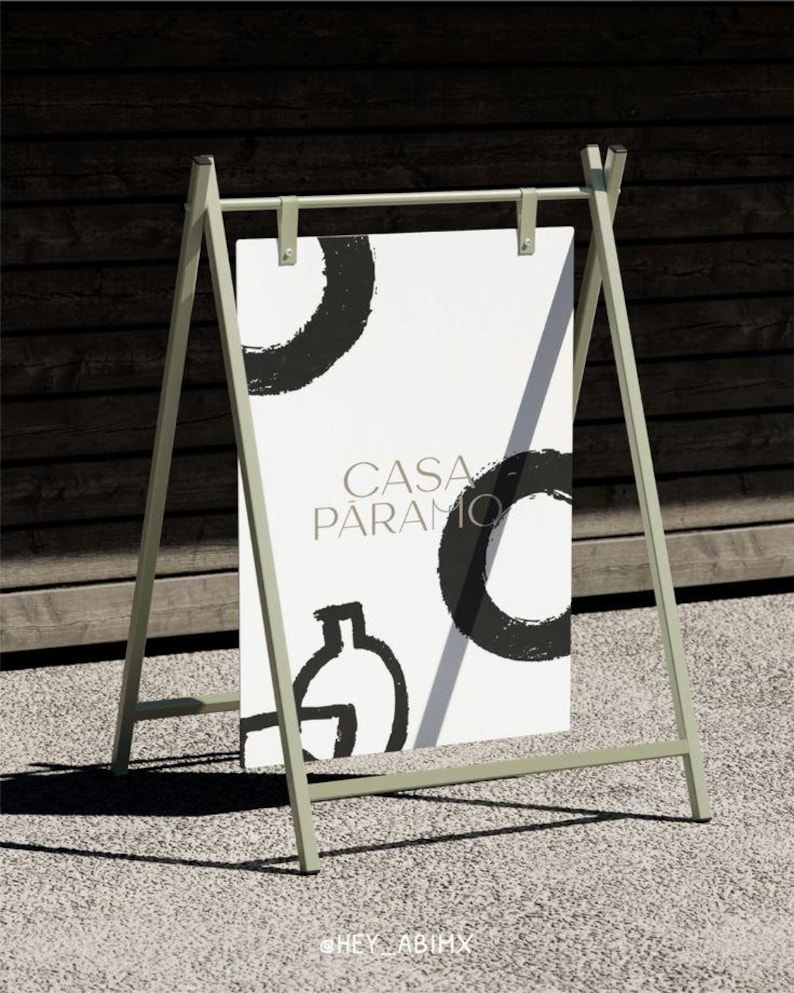 Pu&ograve; includere: Un cavalletto da esterno verde chiaro con un poster bianco. Il poster mostra il testo "CASA PARAMO" e disegni astratti neri. Il cavalletto &egrave; su una superficie grigia, con uno sfondo in legno scuro.