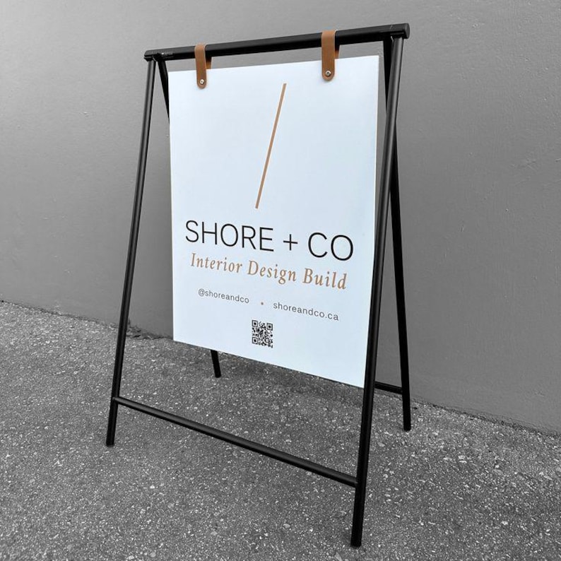 Pu&ograve; includere: Cartello a cavalletto con struttura in metallo nero e cartello bianco. Il cartello presenta il testo "SHORE + CO Interior Design Build" in nero e marrone, con un codice QR e un indirizzo web. Fissato con cinghie in pelle marrone.