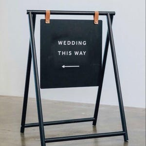 Pu&ograve; includere: Cartello nuziale in metallo nero con la scritta "WEDDING THIS WAY" e una freccia. Il cartello &egrave; attaccato a un telaio in metallo nero con cinghie in pelle marrone. Il cartello &egrave; su un pavimento di cemento.