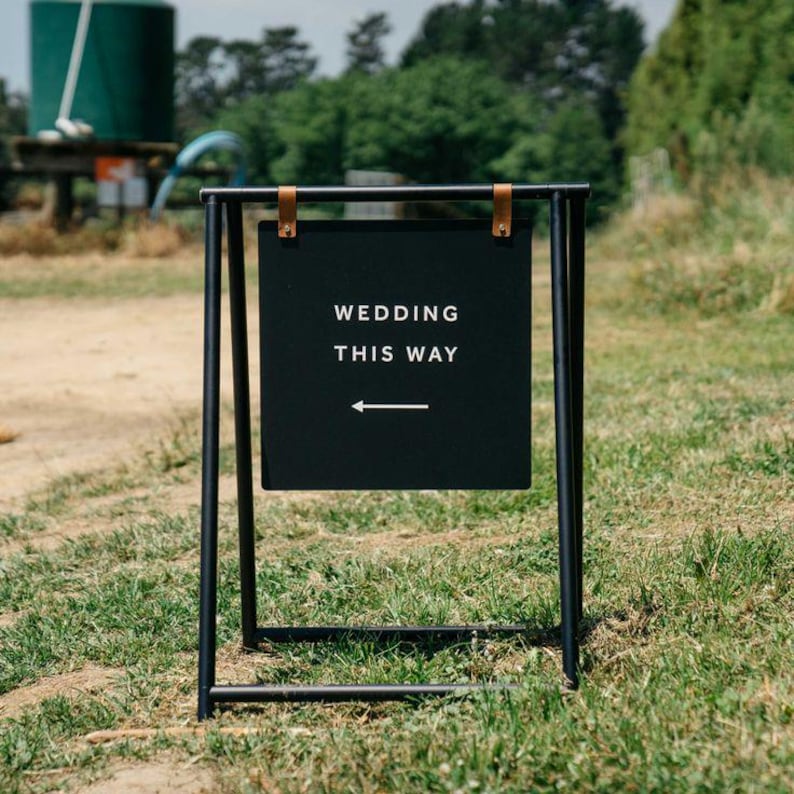 Pu&ograve; includere: Un cartello in metallo nero con le parole "WEDDING THIS WAY" in bianco, con una freccia che punta a sinistra. Il cartello &egrave; sospeso a un telaio in metallo nero con cinghie in pelle. Il cartello &egrave; posto su un'area erbosa.