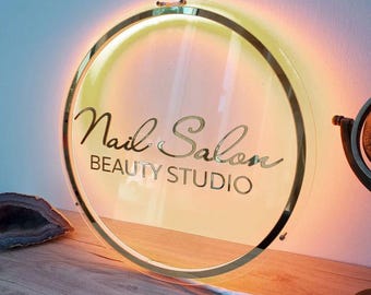 May include: A round, gold-rimmed sign with a white background and the text "Nail Salon Beauty Studio" in gold lettering. The sign is illuminated with warm, yellow light.
