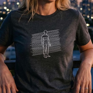 Pode incluir: T-shirt cinzenta escura com um desenho a traço branco de uma figura humana dentro de uma moldura retangular de linhas horizontais. O desenho está centrado no peito. A t-shirt tem mangas curtas e gola redonda.