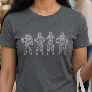 May include: Dark gray t-shirt featuring a white line drawing of four astronauts in space suits. The astronauts are standing in a row, with helmets and various poses. The shirt is made of a soft material.