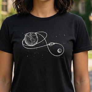 Puede incluir: Camiseta negra con un dibujo lineal blanco de la Tierra, un cohete y la luna. El diseño incluye una trayectoria en bucle que conecta los cuerpos celestes, con estrellas en el fondo. La camiseta es de tejido suave.