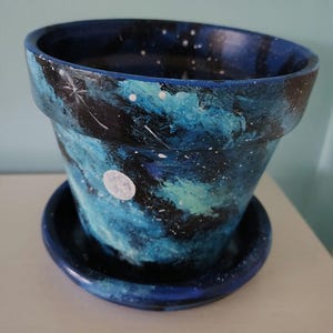 May include: A hand-painted ceramic flower pot with a matching saucer. The pot is painted with a night sky design in shades of blue, black, and white, with a white moon and stars. The pot is approximately 13 cm in diameter.