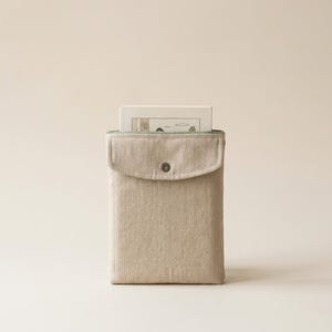 May include: A rectangular beige fabric pouch with a flap closure secured by a single metal snap button. The pouch features a light green interior lining and contains a white rectangular card with a simple line drawing illustration.