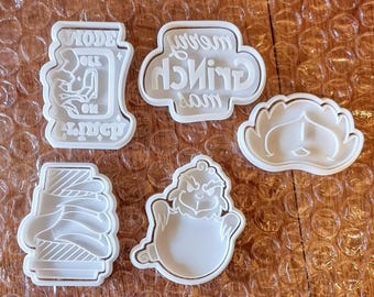May include: A collection of white plastic cookie cutters in various shapes. One cutter has the text "MODELL ON LUNCH". Another says "Merry Grinchmas". Other cutters are shaped like a Grinch face, a stack of presents, and a heart-shaped face.