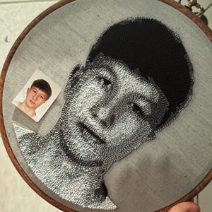 Può includere: Un ritratto ricamato di una persona in bianco e nero, montato in un telaio di legno. Una piccola foto della persona è attaccata al tessuto. Il ricamo presenta ombreggiature e texture dettagliate.