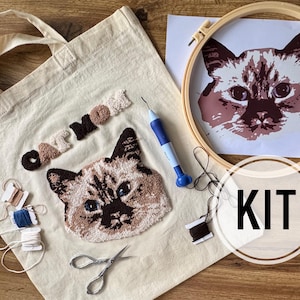 Op de afbeelding: Een knutselpakket met een katten thema, inclusief een beige draagtas, een kattengezicht ontwerp, borduurgaren, een schaar, een punch needle tool en een houten borduurring met een kattengezicht print. Het woord "CAT MOM" is ook inbegrepen.