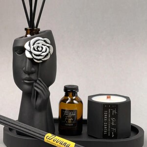 Könnte beinhalten: Ein schwarzes Aromatherapie-Set mit einem gesichtsgeformten Diffusor mit silberner Rose, einer kleinen Flasche, einer Kerze und einem Tablett. Das Set enthält Schilfstäbchen und ein "Dovana"-Etikett. Die Kerze trägt die Aufschrift "TONKA-SANTAL".