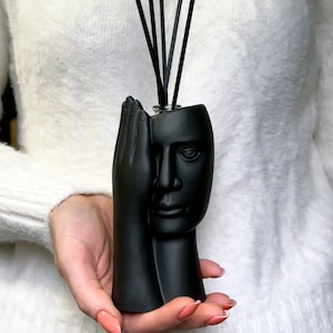 Könnte beinhalten: Ein schwarzer Diffusor mit Gesichts- und Handdesign. Der Diffusor hat ein menschliches Gesicht auf der einen Seite und eine Hand auf der anderen Seite. Schwarze Stäbchen sind in die Oberseite des Diffusors eingesetzt. Der Diffusor wird in einer Hand gehalten.