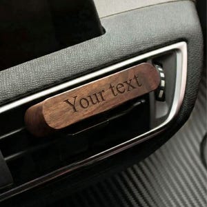 Op de afbeelding: Een houten autoventilatorklem met de gegraveerde tekst "Your text". De klem is rechthoekig en gemaakt van donker hout. Het is in een ventilatieopening van een auto geplaatst, met een zwart dashboard en zilveren afwerking zichtbaar.