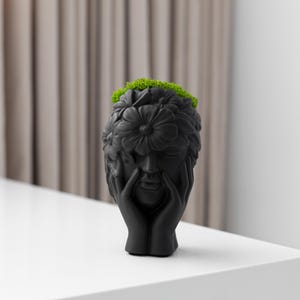 Puede incluir: Una maceta negra con forma de cabeza con una corona floral y manos que sostienen la cara. La maceta está llena de musgo verde. La escultura está sobre una superficie blanca.