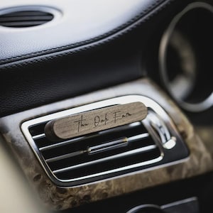 Può includere: Un diffusore di profumo in legno per bocchette d'aria per auto con la scritta "The Oak Faith" in corsivo. Il diffusore è rettangolare e poggia su una bocchetta d'aria argentata. L'interno dell'auto presenta venature del legno e pelle nera.